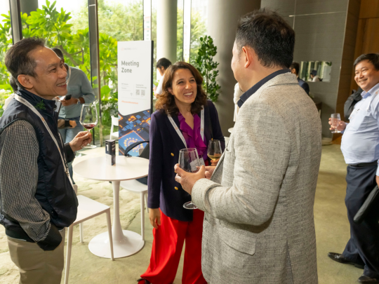 Conference attendees networking over drinks, reflected in a glass wall beside indoor plants.