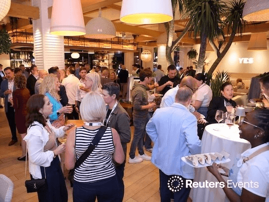 A vibrant, upscale networking reception at a Reuters Events conference, where industry leaders are engaged in conversation, showcasing the high-level professional community and peer-to-peer connection that defines the event experience.