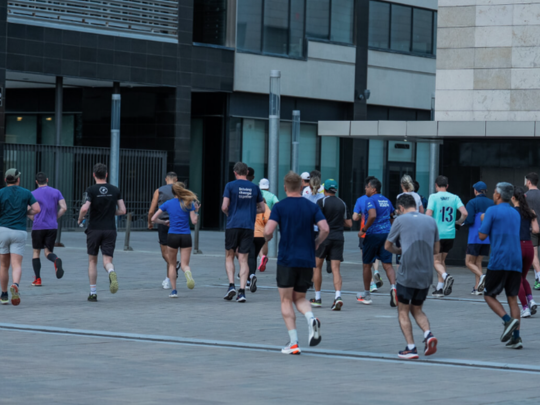 Approximately 25 runners of varying ages participate in an outdoor 5K fun run at a pharma conference, jogging across a modern urban plaza.