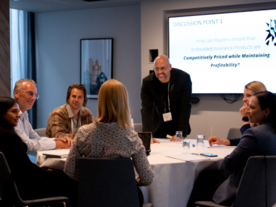 Six conference attendees take part in a roundtable discussion as a facilitator stands beside a screen displaying a discussion prompt.