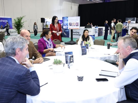 Attendees gather around a round table for Day 0 networking at Energy LIVE, with a Meeting Concierge sign and expo booths visible in the background.