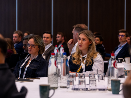A room full of professionals attentively listening to a presentation at the Supply Chain Europe conference, seated at white-clothed tables with water bottles.