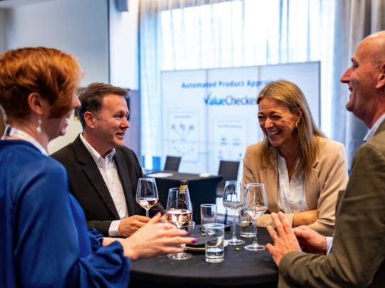 Four insurance professionals laughing and networking over drinks at a standing table during a conference evening reception, with a ValueChecker display visible in the background.