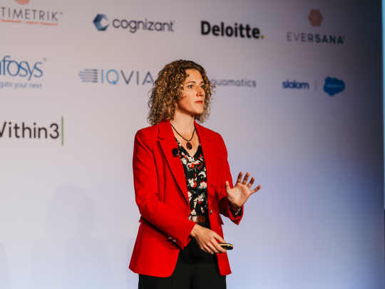 Woman in a red blazer presenting on a conference stage in front of sponsor logos.