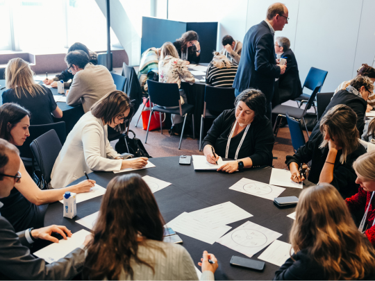 Attendees participate in small-group workshop activities at tables, writing on worksheets and discussing ideas.