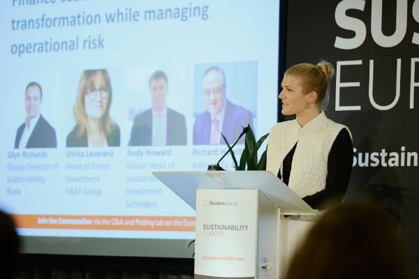 A speaker delivering opening remarks at the podium with speaker profile slides displayed in the background at Sustainability Europe.