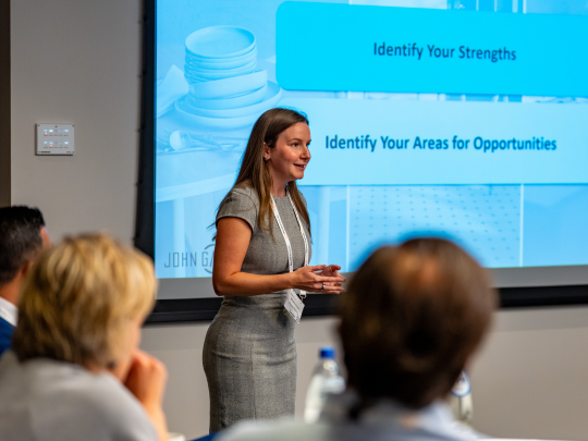 A female speaker presenting to a seated audience at Supply Chain Europe, with a projected slide reading 'Identify Your Strengths' and 'Identify Your Areas for Opportunities' displayed behind her.