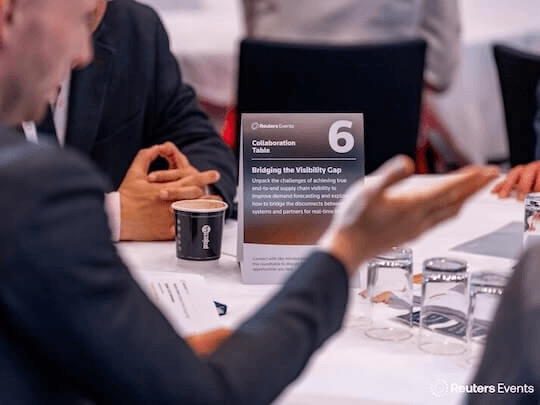 A close-up view of a 'Collaboration Table' centerpiece at a Reuters Events conference, titled 'Bridging the Visibility Gap.' The image captures attendees in focused, collaborative discussion, highlighting the interactive, problem-solving nature of the sessions.