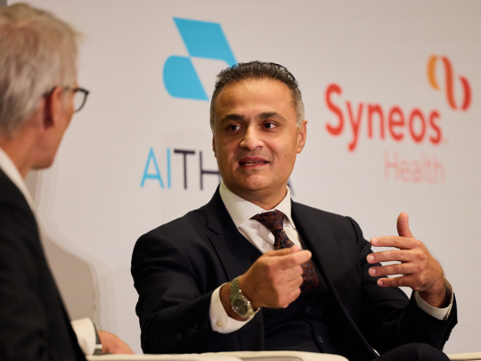 Man in a dark suit speaking during a seated conference discussion in front of AIT and Syneos Health logos.