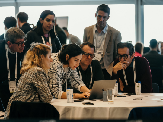 Conference attendees gather around a table, reviewing a laptop during a collaborative roundtable discussion.