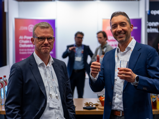Two male attendees smiling and posing for a photo in the exhibition hall at Supply Chain Europe, with event sponsor banners and other networking attendees visible in the background.