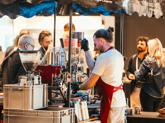 A barista preparing fresh coffee at a mobile station during a busy networking break at a Reuters Events conference, with professional attendees engaged in conversation in the background, highlighting the vibrant social atmosphere of the event.