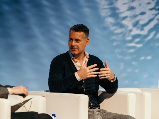 Man speaking from a white armchair during a panel discussion, gesturing with both hands.