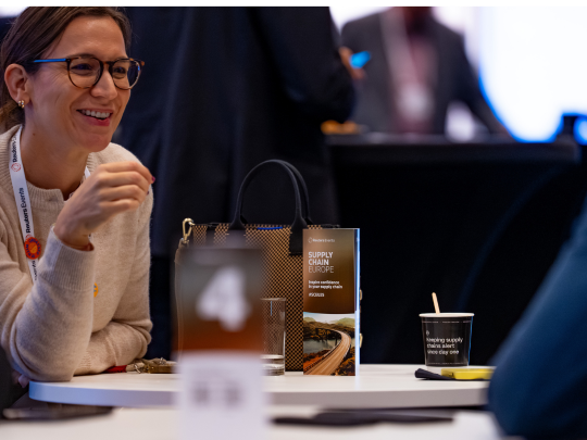 A smiling attendee at the Supply Chain Europe conference, with a branded tote bag and Supply Chain Europe brochure visible on the table.