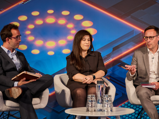 Panel discussion with three speakers seated on stage against a digital technology backdrop at a Reuters Events conference.