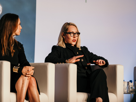 Woman wearing glasses speaking from a white armchair on a conference panel, with another panelist beside her.