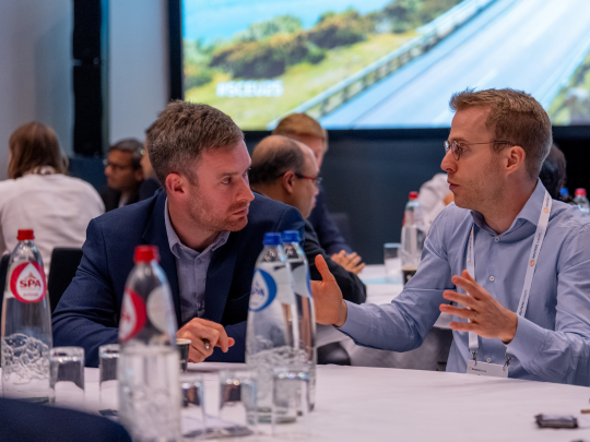 Two male delegates engaged in a focused one-to-one conversation at a roundtable session during Supply Chain Europe, with water bottles and other attendees visible in the background.