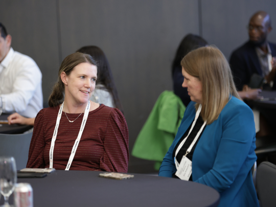 Two Total Health attendees wearing event badges talk at a round table during a conference break