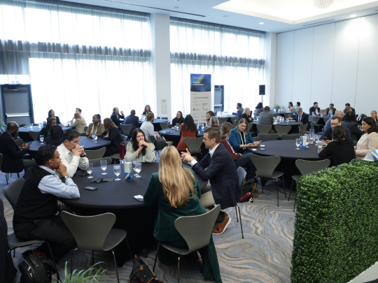 Attendees network in small groups at round tables during Total Health conference, in a bright venue with large windows and a green wall feature.