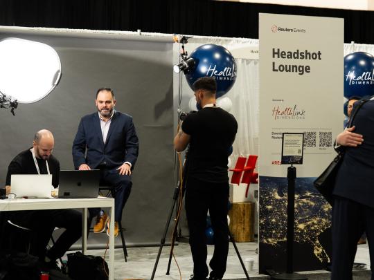 Male executive on bar stool posing for a headshot with camera man and assistant at the headshot lounge at Reuters Events Pharma USA 2026