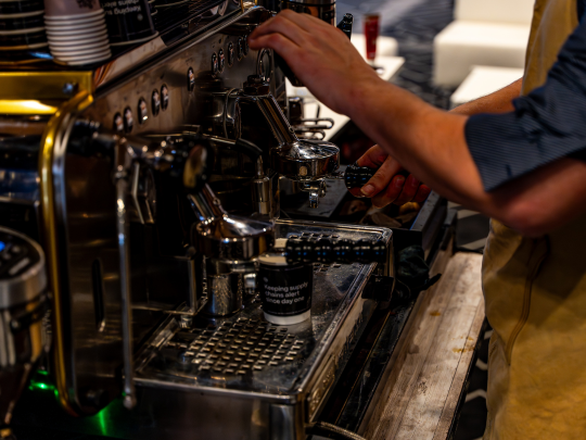 A barista preparing coffee on a professional espresso machine at the Supply Chain Europe conference, with a branded cup in the foreground.