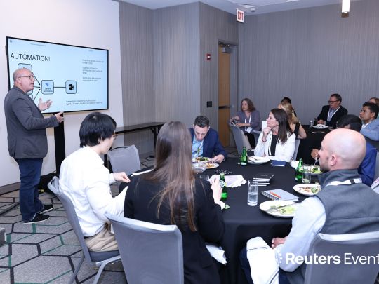 An interactive roundtable workshop at The Future of Insurance 2026. A presenter stands next to a screen showing an automation workflow diagram, addressing a small, focused group of insurance executives seated around a table.