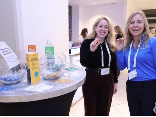 Two women smiling and holding product samples at a branded exhibition stand during an insurance industry conference or trade show.