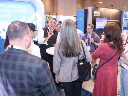 A crowd of insurance industry attendees gathered around an exhibition stand on the expo floor, engaged in lively conversation with a presenter.