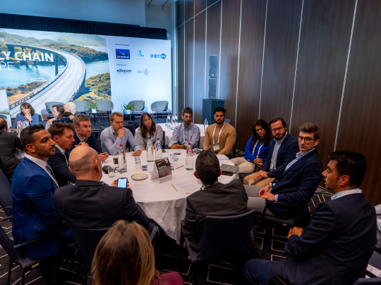 Supply Chain Europe attendees take part in a roundtable discussion at a conference table, with the event stage and sponsor display in the background.