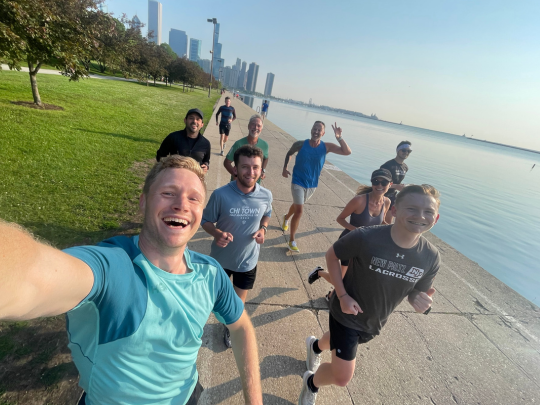 A group of runners take a selfie during a waterfront fun run, with a skyline visible in the background.