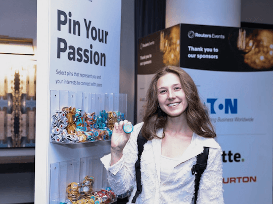 A smiling attendee at a Reuters Events conference holding a pin at the 'Pin Your Passion' interactive networking station, designed to help delegates connect with peers based on shared professional interests.