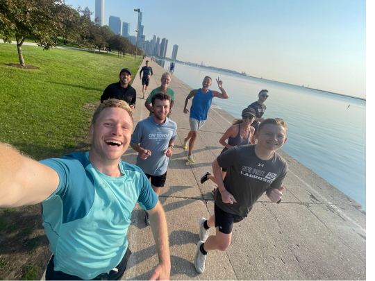 A group of runners take a selfie while jogging along a waterfront path with a city skyline in the background.
