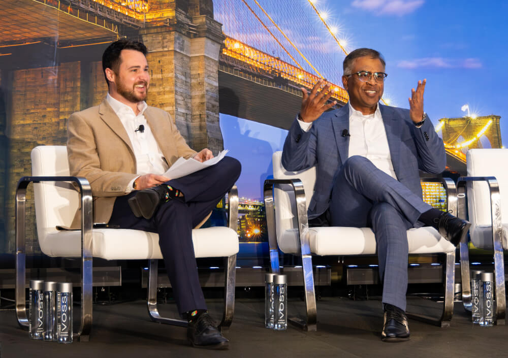 Two speakers engaged in a dynamic discussion on stage at a Reuters Events conference, seated in modern white armchairs against a large backdrop of the Brooklyn Bridge.